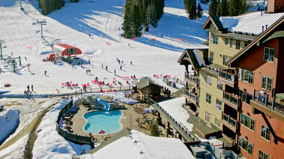 The image features the energetic base of a mountain resort, with snow-covered ski slopes, a vibrant pool area, a ski lift in action, and the welcoming exterior of the lodge. Families and groups appear to be enjoying various winter and après-ski activities.