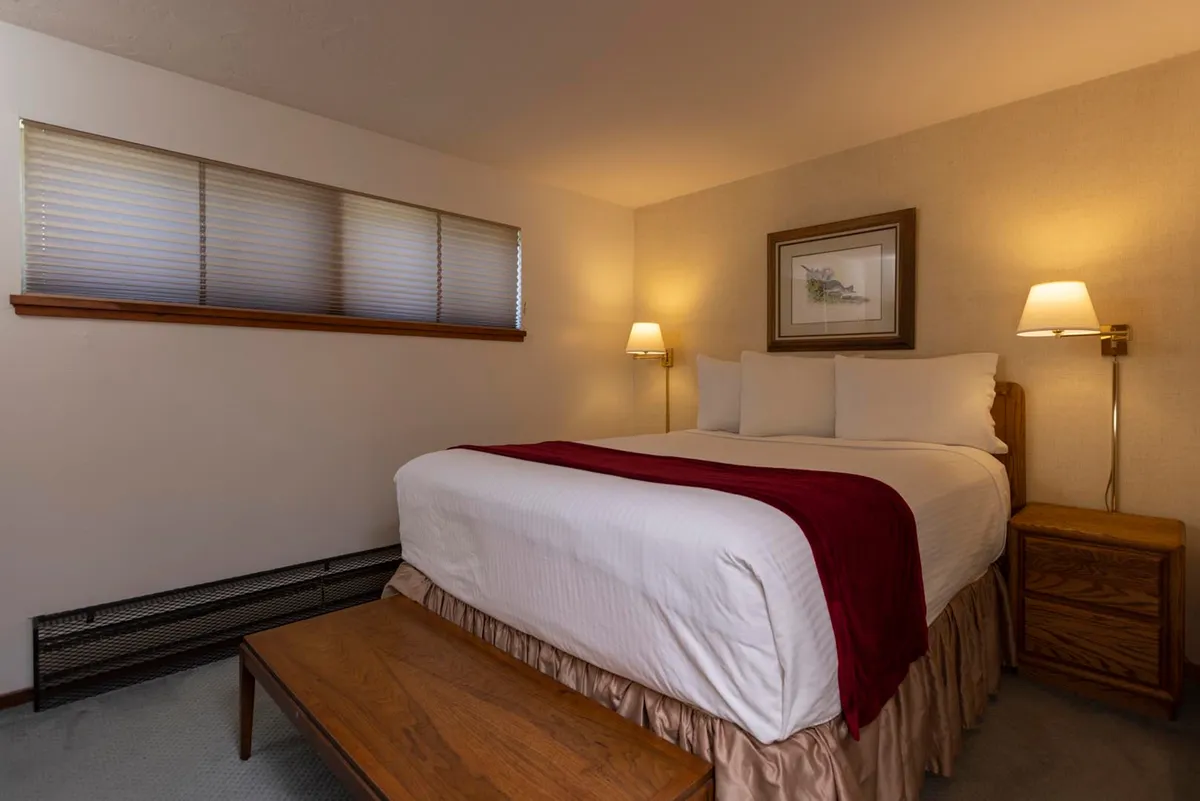 A cozy master bedroom with a plush bed, soft white linens, and warm lighting from table lamps. The accompanying nightstand and tasteful wall art create a restful, welcoming atmosphere, perfect for winding down after a day in the mountains.