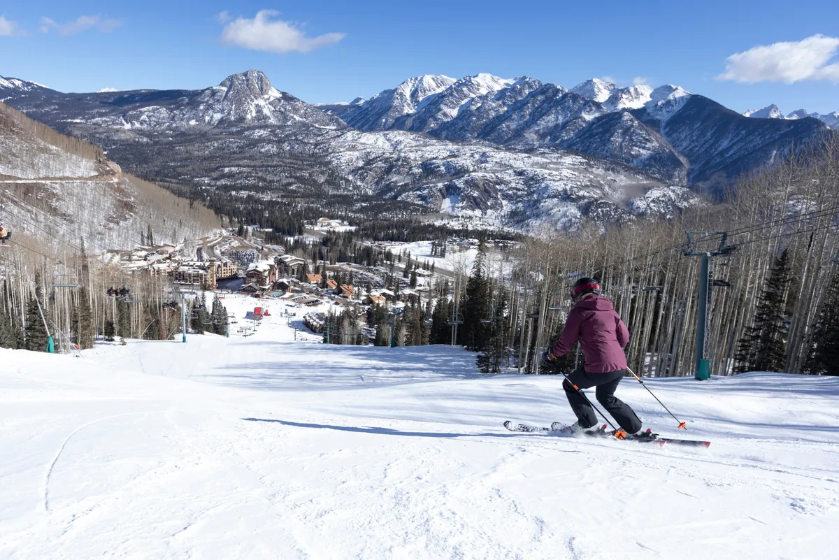 A dynamic scene featuring a skier descending a sunlit slope with a distant mountain range and the ski resort village visible below. The image conveys energy, excitement, and the thrill of mountain sports, with a sense of community from the resort buildings.