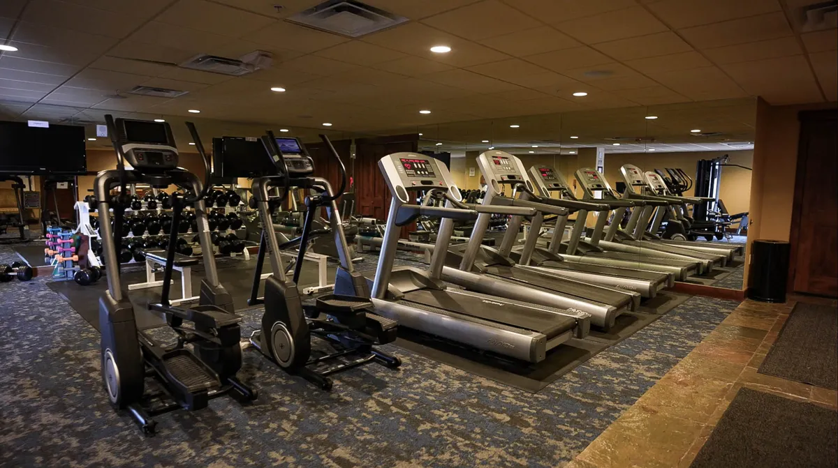 This image showcases a modern fitness center equipped with elliptical machines, treadmills, a dumbbell rack, and a mirrored wall. The environment is bright and invigorating, inviting guests to pursue fitness goals or warm up before heading outdoors. The space suggests both individual and group exercise options.