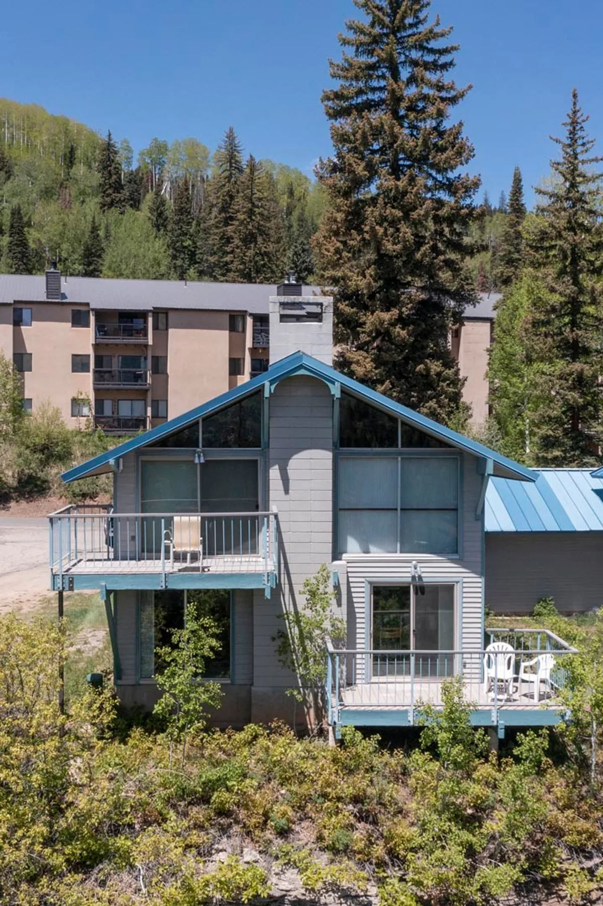 This peaceful exterior view showcases the house with its notable blue roof and inviting balcony, nestled among lush, mature trees. The setting encourages relaxation on the balcony and exploration of the surrounding natural environment, suggesting the proximity of trails and opportunities for family gatherings.