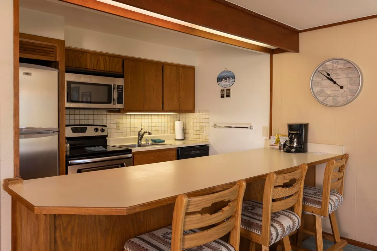 The focal point is a functional kitchen island with bar stools, stainless steel appliances, and wooden cabinetry. The area exudes warmth and practicality, inviting both cooking and socializing. Decorative elements like a wall clock and art add a personal touch.