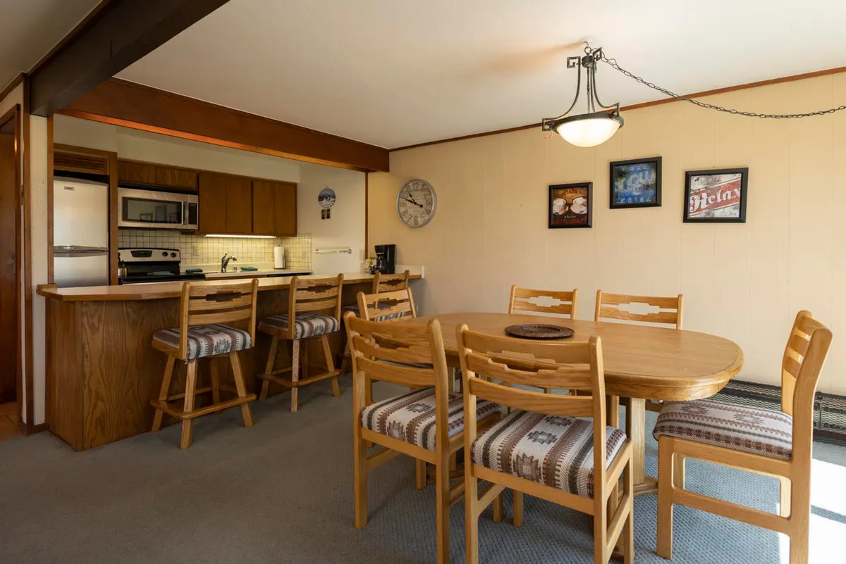 A bright, inviting scene with a focus on the dining table, bar seating, and a kitchen area in the background. Decorative wall art adds character, while the table is set for a meal or gathering.