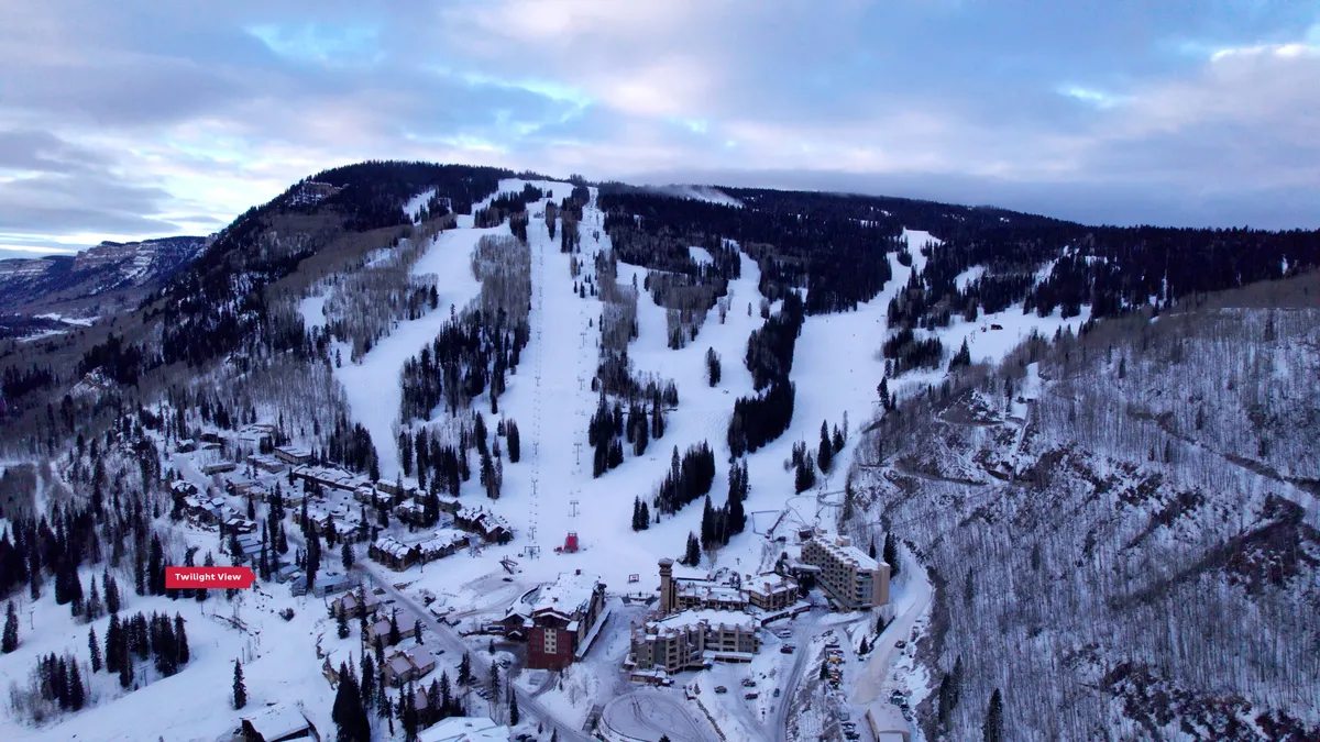 A sweeping view of the snow-covered ski slopes, dramatic mountains in the background, resort buildings nestled below, and a scatter of pine trees. The scene is tranquil, with hues of sunset casting a golden glow across the landscape, emphasizing the proximity to all-season mountain adventure.