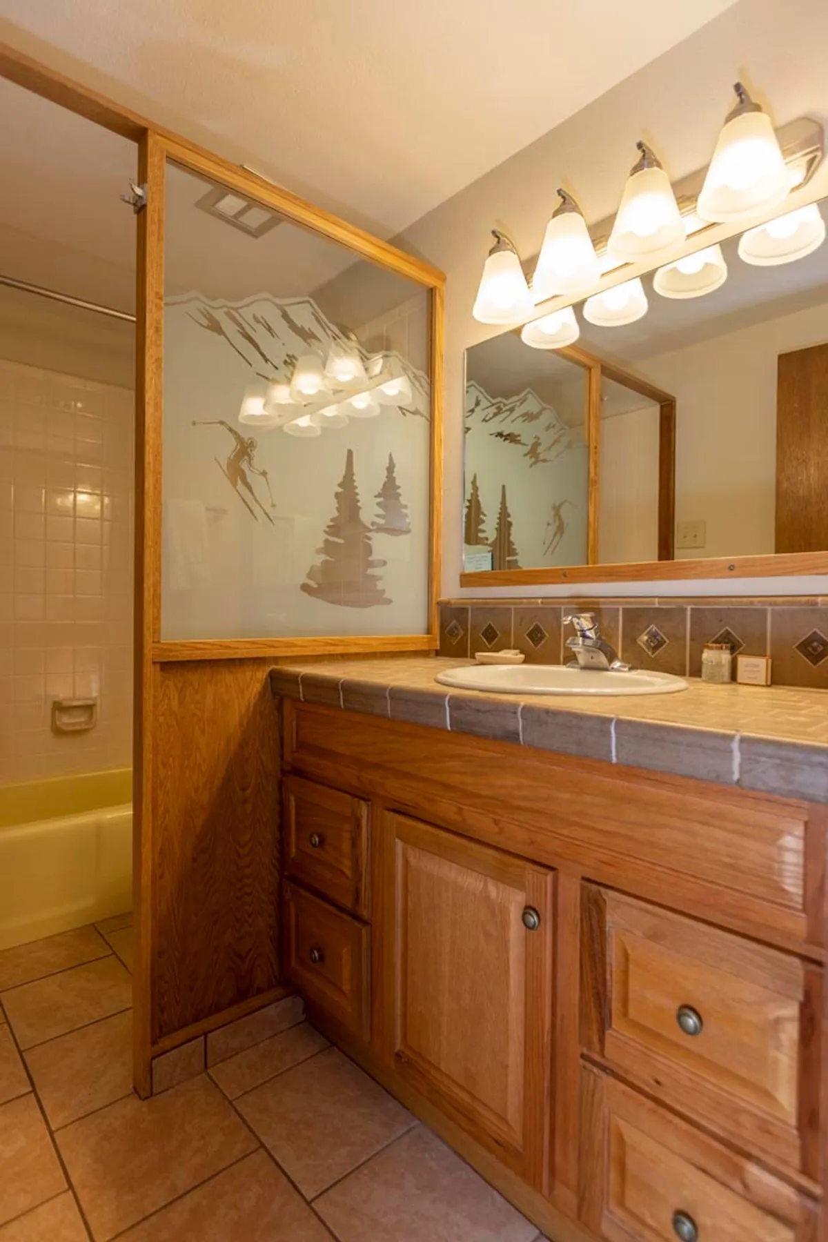 A modern, serene bathroom with a wooden vanity, decorative glass partition adorned with mountain artwork, and a luxurious bathtub in the corner. The scene evokes both morning routines and evening relaxation, reinforcing the retreat's spa-like amenities.