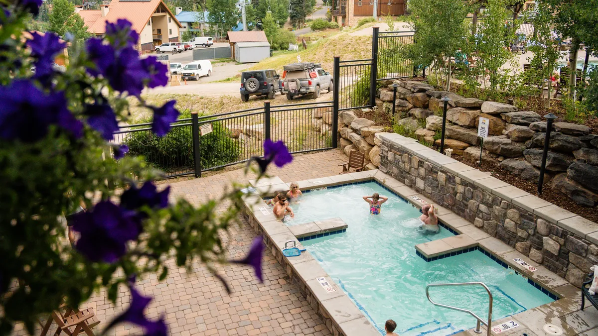 A sunlit pool and hot tub are surrounded by vibrant purple flowers and natural rock walls, with expansive views of the surrounding landscape. The image radiates a sense of leisure and family fun in the warmer months, with space for swimming, relaxing, and socializing outdoors.