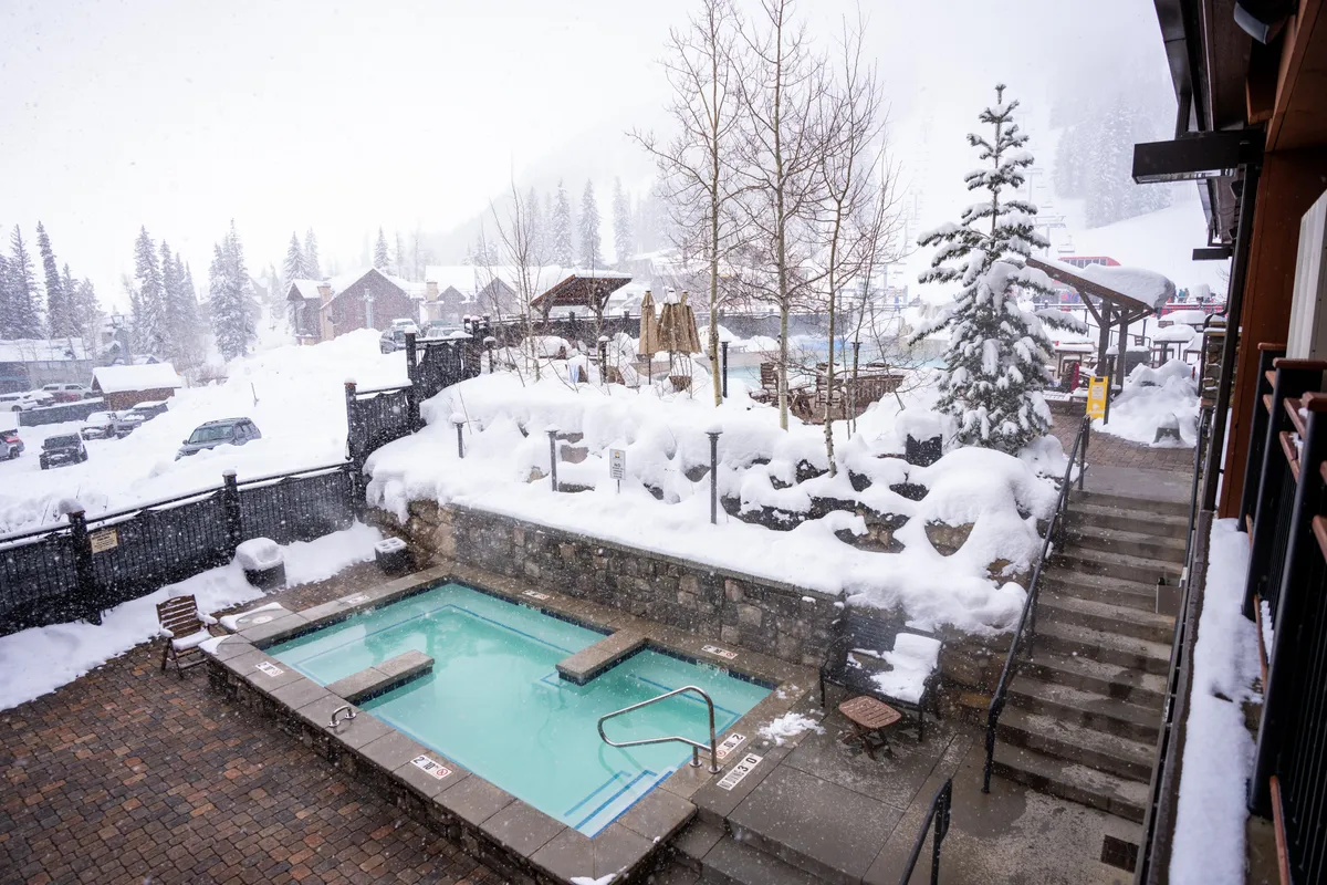 An inviting outdoor hot tub surrounded by snow-covered trees, with a stone wall and steps leading up, creating a tranquil and cozy scene. The steam rising from the hot tub contrasts with the crisp winter landscape, evoking the comfort and relaxation at the end of a day spent in the mountains.